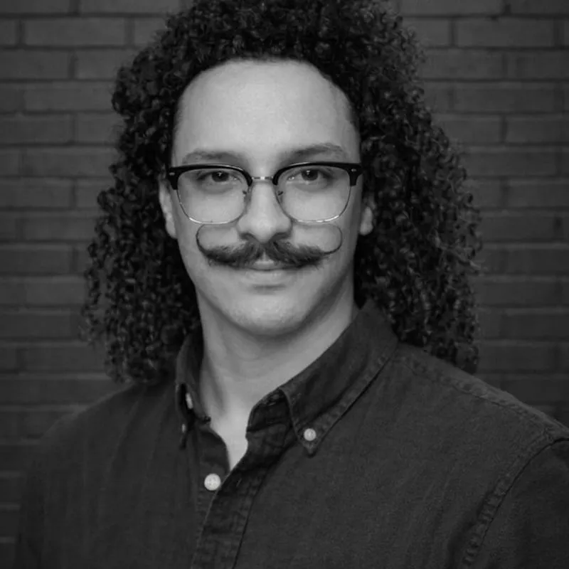 Man with long curly dark hair and handlebar mustache wearing glasses and a dark button-up shirt, professional headshot portrait