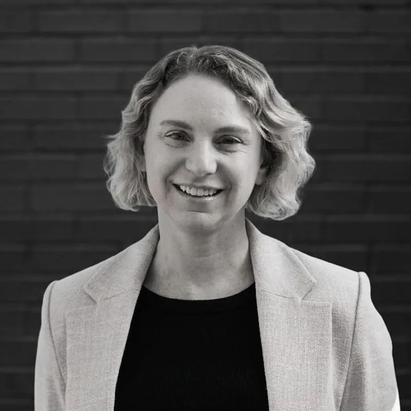 Professional woman with short wavy blonde hair wearing light gray blazer over black top, smiling confidently against a dark background